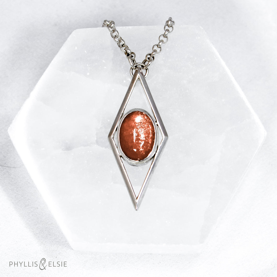 A rich orange oval Sunstone is centered in a diamond frame of polished silver. Beveled edges catch the light and highlight the stone floating in the center.  Details  Solid sterling silver, partially recycled  Sunstone  Pendant: 17mm x 41mm  18