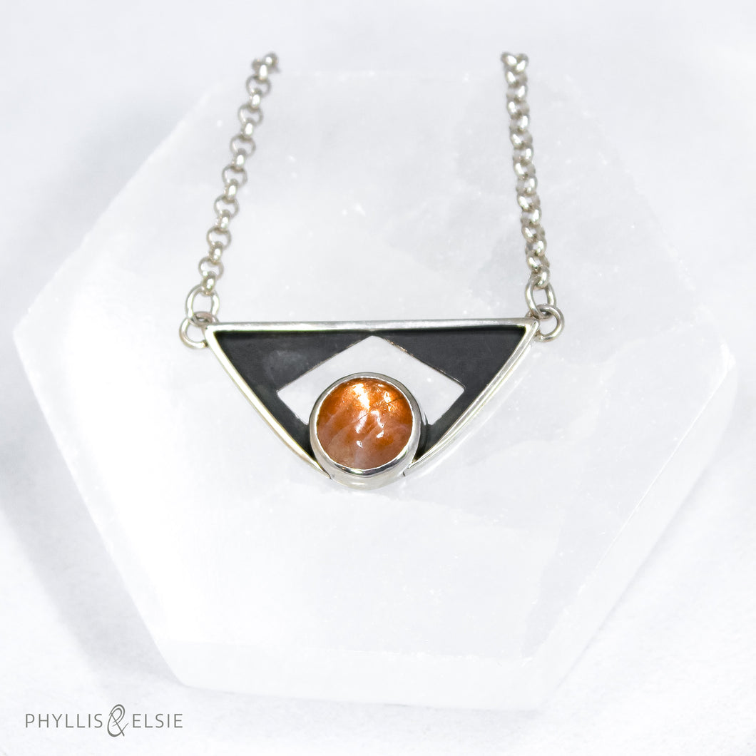 A glowing Sunstone set in a striking patinated shadowbox with a geometric window. The crisp lines of this bold pendant are balanced with a heavy silver rolo chain.  Details:  Solid sterling silver, partially recycled  Sunstone  Pendant: 35mm x 17mm  18
