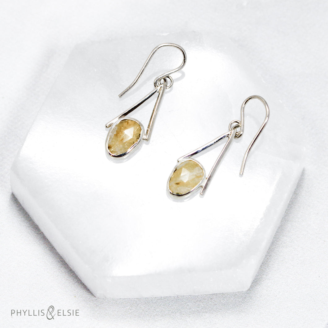 Rosecut Quartz with golden hued crystalline inclusions are suspended in an airy, asymmetrical frame  Details:  Solid sterling silver, partially recycled  Rutilated Quartz  Earring faces: 11mm x 23mm  Sterling silver ear hooks