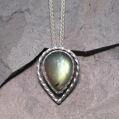 This Stella necklace features a glowing, smooth sage-green Labradorite teardop cabochon that shows glimpses of peach and aqua in bright light. Surrounded with a slightly-offset twisted halo, this pendant stands out despite its small size and soft colors.